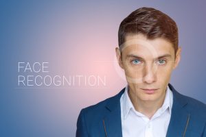 Es ist ein Mann zu, sehen dessen Auge durch eine transparenten Zielscheibe fokussiert wird. Daneben ist der Schriftzug "Face Recognition" geschrieben. / Gesichtserkennung